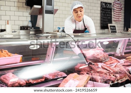 Positive young butcher shop saleswoman arranging meat products on display case, showing piece of fresh raw veal tenderloin Royalty-Free Stock Photo #2419526659