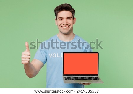 Young IT man wear blue t-shirt white title volunteer hold use work on blank screen laptop pc computer show thumb up isolated on plain green background. Voluntary free work help charity grace concept