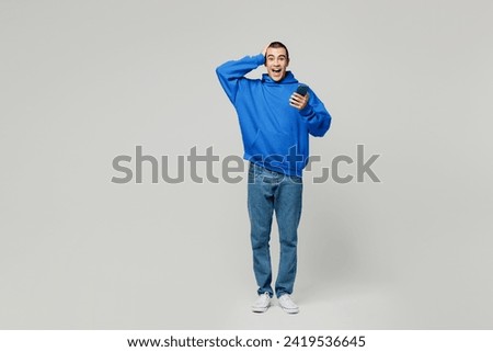 Full body surprised amazed young middle eastern man he wear blue hoody casual clothes hold head hand use mobile cell phone isolated on plain solid white background studio portrait. Lifestyle concept