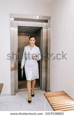 Vertical portrait of female doctor in white coat getting off elevator and walking down hospital corridor holding clipboard in hand. Young woman practitioner walking along hallway holding folder