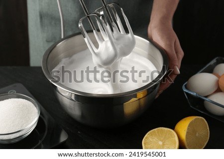 Woman making whipped cream with hand mixer at black table, closeup