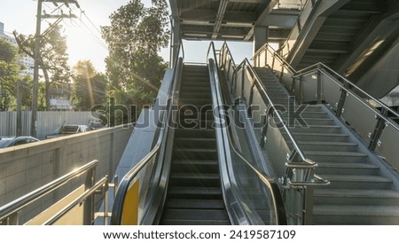 Photograph of an urban location, an escalator flanked by cement stairs. Handrails made of steel and glass, under roofs, public areas Outside the wall was a narrow alley, cars parked close together.