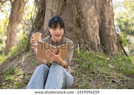 A happy and pretty Asian woman in casual clothes is sipping coffee and reading a book under a tree in a garden. people and leisure activity concepts