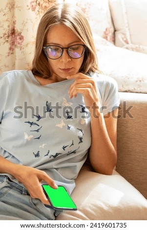 Young girl in eyeglasses sitting at couch at home holding smartphone with green screen in her hands looking at it. Online pastime, shopping, communication, leisure. Chroma key mock up. Vertical format