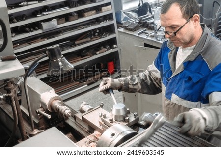 Portrait of professional turner at work on lathe in workshop. 50-55 year old turner in overalls and glasses turns part in workshop on machine. Photography authentic work process.