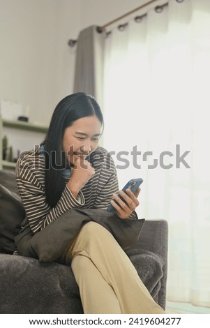 Smiling young woman chatting with friends on mobile phone, sitting on couch at home