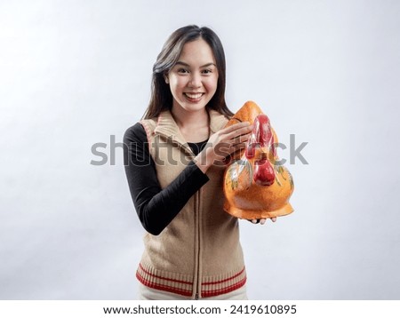 Portrait of an Indonesian Asian woman, wearing a light brown sweater, posing while holding a chicken-shaped piggy bank, isolated against a white background.