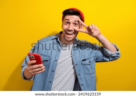 Photo of positive good mood man dressed jeans shirt showing v-sign cover eye texting modern gadget isolated yellow color background