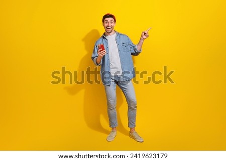 Full length photo of excited funky man dressed jeans shirt typing modern gadget pointing empty space isolated yellow color background