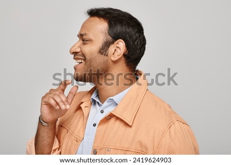 joyful indian man in vibrant orange jacket smiling happily and looking away on gray backdrop