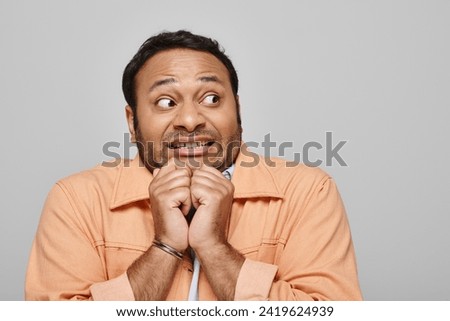 scared indian man in bright orange jacket posing frightened and looking away on gray backdrop
