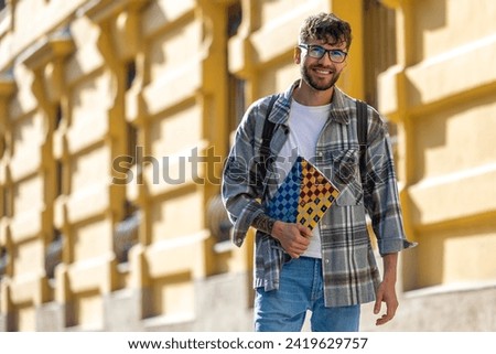 Male student in glasses holding textbooks and smiling while standing at university building. Royalty-Free Stock Photo #2419629757