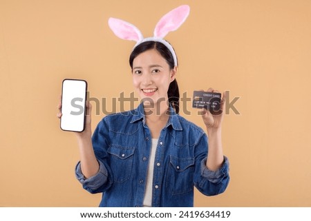 Portrait asian young woman wearing denim clothes and bunny rabbit ears and holding smartphone and credit card isolated on beige background. Shopping Happy Easter concept.