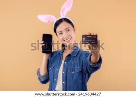 Portrait asian young woman wearing denim clothes and bunny rabbit ears and holding smartphone and credit card isolated on beige background. Shopping Happy Easter concept.