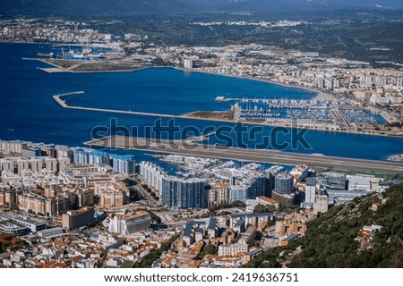 Gibraltar, Britain - January 24, 2024 - An aerial view of a densely built coastal city with a marina, an airport runway near the sea, industrial areas, and mountains in the background.