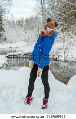 Full-length portrait of a young Caucasian red hairy woman in the winter snowy forest. Young woman wearing sport glasses, blue jacket and black tights looking over the shoulder into the camera