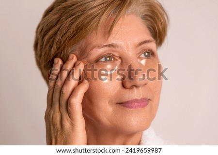 elder woman doing facial care daily routine procedure.granny applying serum,cream,remove makeup with cotton disk.female with towel on head and mask sheet on face is drinking coffee,wellbeing.