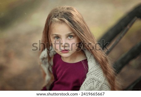A beautiful little girl with long blond hair in the forest is sad. The little girl looks calmly into the camera. Portrait of a girl with long blond hair blowing in the wind. 