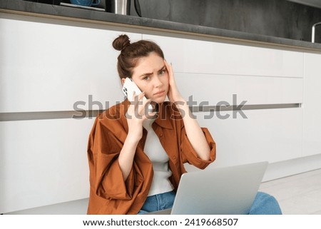 Image of woman with concerned, upset face, sits on floor with laptop, calling on phone, listens bad news.