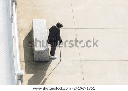 Elderly woman with a crutch next to a bench on a street looking at the ground. Copy space. High angle view Royalty-Free Stock Photo #2419691951