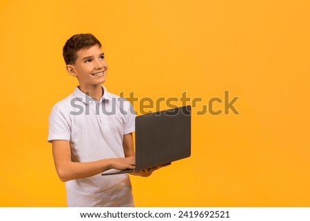 Thoughtful teenage boy holding an open laptop, gazing upwards with inspired expression, smiling male kid dressed in white polo shirt standing against yellow studio background, copy space