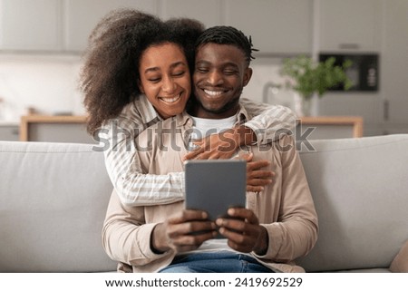 Cheerful African American Couple Using Digital Tablet Browsing Internet, Woman Hugs Husband While He Holding Computer Gadget, Websurfing And Exploring Online Offers At Home Interior Royalty-Free Stock Photo #2419692529