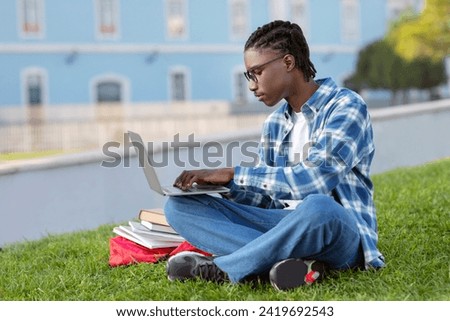 Focused black guy engages in e-learning on his laptop, typing and websurfing for academic project, seated on lawn at campus park outdoor, side view shot of student with computer Royalty-Free Stock Photo #2419692543