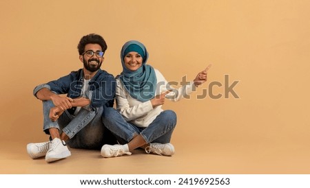 Check This. Young Arabic Spouses Pointing Aside At Copy Space, Muslim Man And Woman In Hijab Showing Free Place For Your Advertisement Or Offer, Sitting Together On Beige Background, Panorama Royalty-Free Stock Photo #2419692563