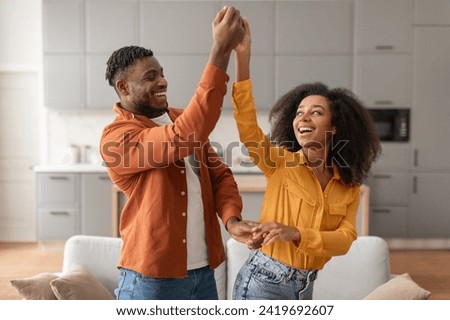 Cheerful millennial black spouses having fun together dancing to music in modern kitchen interior, smiling to each other. Couple having date or party. Relationship and romance concept