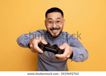 Excited asian guy playing video games with joystick over yellow studio background, happy millennial male gamer looking at camera with excitement, enjoying virtual entertainment, copy space