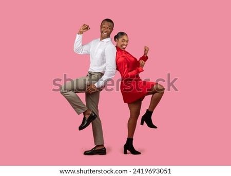 Energetic black man in white shirt and woman in red dress, both with fists raised in powerful victory pose, standing on one leg against pink background