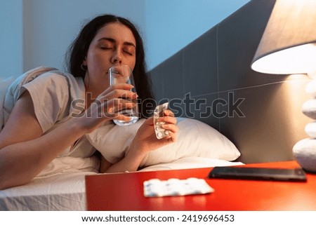 Caucasian adult sleeping woman lies in bed and drink water and holds medicines. Concept of insomnia, sleep disorders and headache.