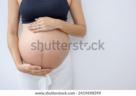 Close-up photo of pregnant woman with pregnancy holding her large belly standing against the wall in a white room at home.