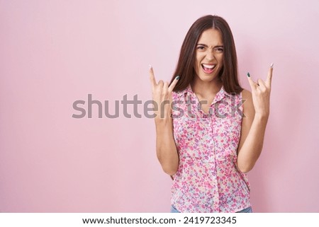 Young hispanic woman with long hair standing over pink background shouting with crazy expression doing rock symbol with hands up. music star. heavy concept. 
