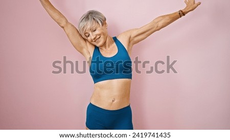 A joyful mature woman stretches in athletic wear against a pink background, depicting wellness and vitality.