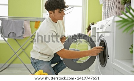 Smiling young hispanic teenager enjoying doing laundry, confidently inserting detergent bag into washing machine in laundry room