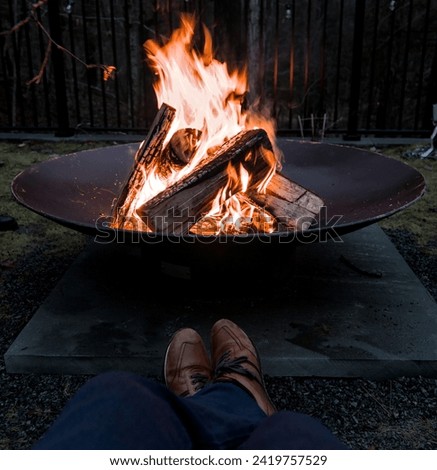 man sitting in front of fire pit (jeans, pants, shoes) looking down at burning campfire (winter outdoor activity) Royalty-Free Stock Photo #2419757529