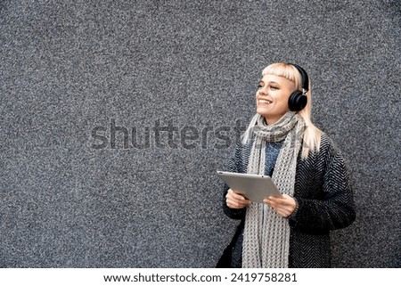 Happy hippie girl browsing a tablet and listening to the music with headphones leaning on a wall in the street. Hipster woman online working while waiting colleague listen to music on wireless headset Royalty-Free Stock Photo #2419758281