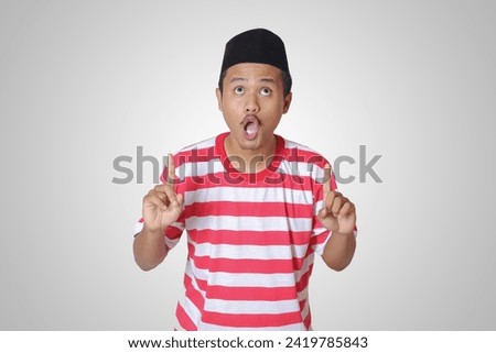 Portrait of attractive Asian man in red white stripe t-shirt with skullcap pointing with two hands and fingers to the up side. Advertising concept. Isolated image on gray background