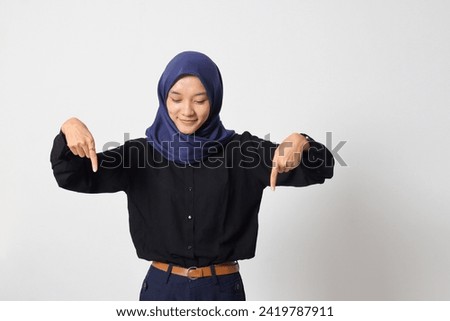 Portrait of excited Asian hijab woman in casual shirt promoting product, pointing finger down. Advertising concept. Isolated image on white background