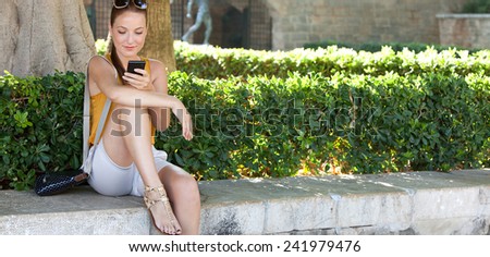 Young and beautiful tourist woman sitting in a green park holding and using her smartphone to network while visiting a destination city on holiday. Travel and lifestyle technology.