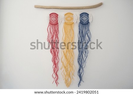 Three macramé circles in Romanian flag colors-red, yellow, and blue-hang elegantly on a wooden branch. Suspended against a white backdrop, the fringed ends form a striking composition.
