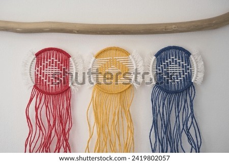 Three macramé circles in Romanian flag colors-red, yellow, and blue-hang elegantly on a wooden branch. Closeup against a white backdrop