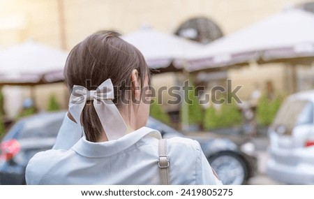 A fashionable woman with a stylish hair bow confidently strolls down a city street. She exudes elegance in her white blazer and black pants while checking her phone amid the urban buzz and energy.