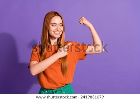 Portrait of positive champion girl wear orange t shirt pointing finger her huge biceps after trainings isolated on violet color background
