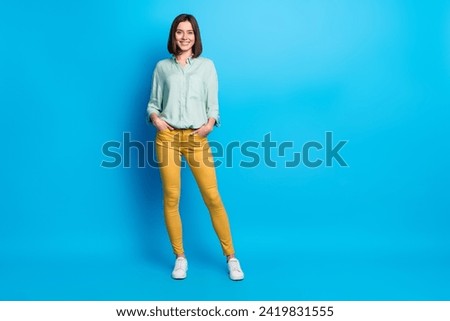 Full size photo of positive cool pretty girl with stylish hair wear teal shirt standing arms in pockets isolated on blue color background