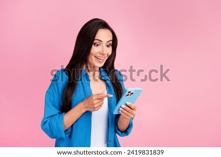 Portrait of excited impressed woman with long hair wear stylish clothes look indicating at smartphone isolated on pink color background
