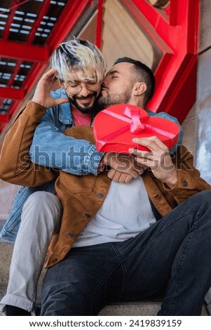 Young adult couple gay sitting outside celebrating valentine's day, his boyfriend is reciving a gift with heart shape and he is kissing him with emotion and affection for the surprise. Royalty-Free Stock Photo #2419839159