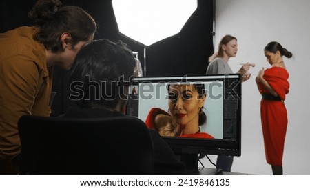 Backstage of model and professional team in the studio. Editor and photographer talking checking photos looking at monitor, assistant fixes makeup.