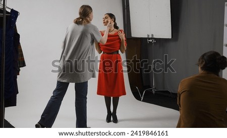 Backstage of model and professional team in the studio. Female assistant fixes makeup for model in red dress, photographer taking photographs.